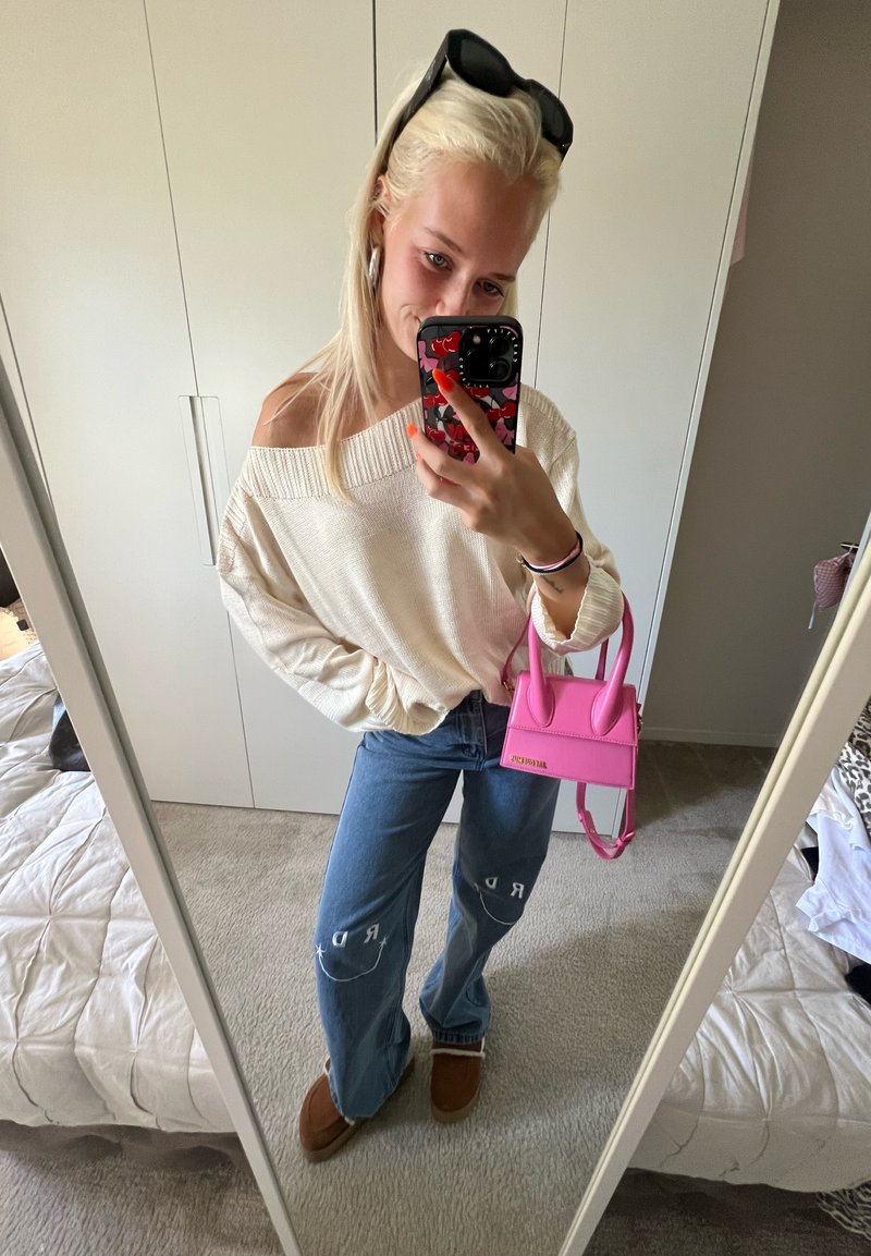 Oversized μπεζ πουλόβερ, ανοιχτού μπλε παντελόνι με φαρδιά πόδια και λεπτομέρεια λογότυπου, καφέ παντόφλες και μικρή ροζ τσάντα χεριού με δομητές λαβές.