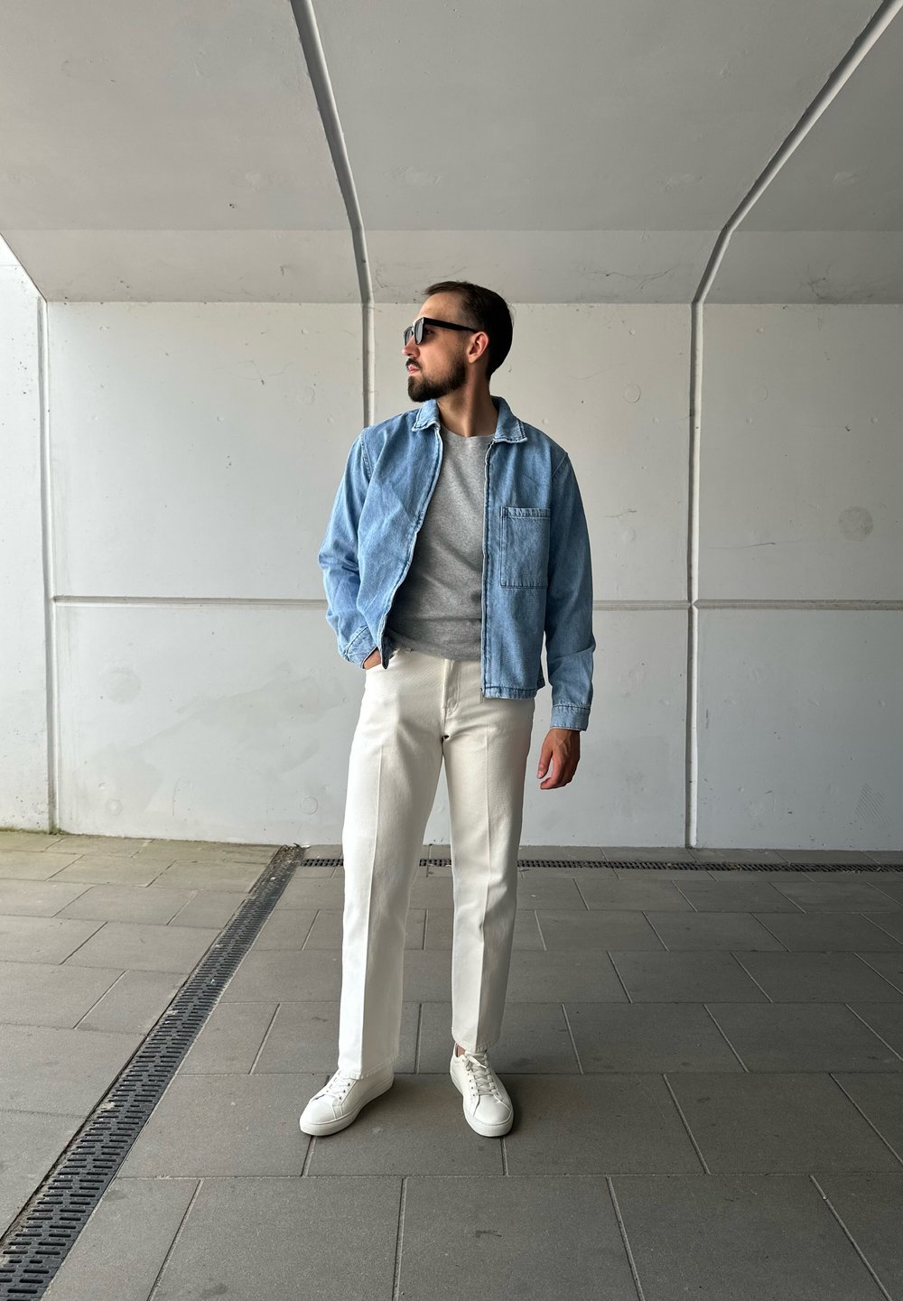 Chaqueta de mezclilla azul claro sobre un suéter gris, pantalones rectos blancos y zapatillas blancas. Unas gafas de sol negras y brillantes completan el look.