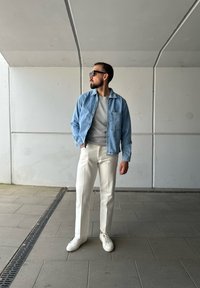 Chaqueta de mezclilla azul claro sobre un suéter gris, pantalones rectos blancos y zapatillas blancas. Unas gafas de sol negras y brillantes completan el look.