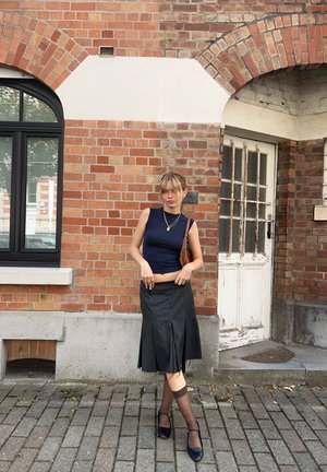 Top sin mangas de color azul marino, falda negra con pliegues, bolso marrón de hombro. El fondo presenta una pared de ladrillos y una puerta de madera con cristales.