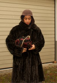 Manteau en fausse fourrure noire avec un col montant, présentant une texture matelassée. Tenant un sac à main à motif floral avec une bandoulière en chaîne dorée.