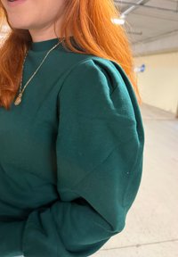 Pull-over vert avec épaules bouffantes, confectionné dans un tissu doux. Présente une finition texturée et des poignets côtelés. Exposé dans un environnement intérieur bien éclairé.