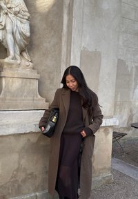 Manteau marron oversize par-dessus un pull sombre, associé à un sac noir. En arrière-plan, une statue en pierre et un mur texturé.