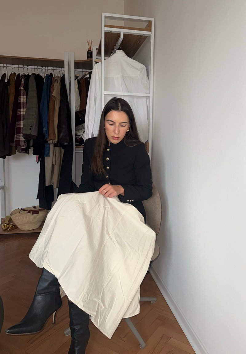 Schwarze Blazer mit goldenen Knöpfen, cremefarbener Stoff der auf dem Schoß drapiert ist, schwarze High-Heels, und ein Hintergrund aus hängenden Kleidungsstücken.