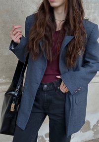Blazer cinza com lapelas largas, tecido texturizado e detalhes de punhos com botões, combinado com uma blusa bordô e jeans pretos de cintura alta. Bolsa tote preta.