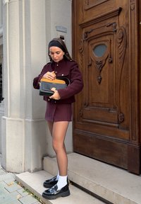 Jachetă din lână bordeaux cu butoane aurii, pantaloni scurți bordeaux asortați, geantă de umăr neagră cu un accent auriu și loafers negri chunky cu șosete albe.