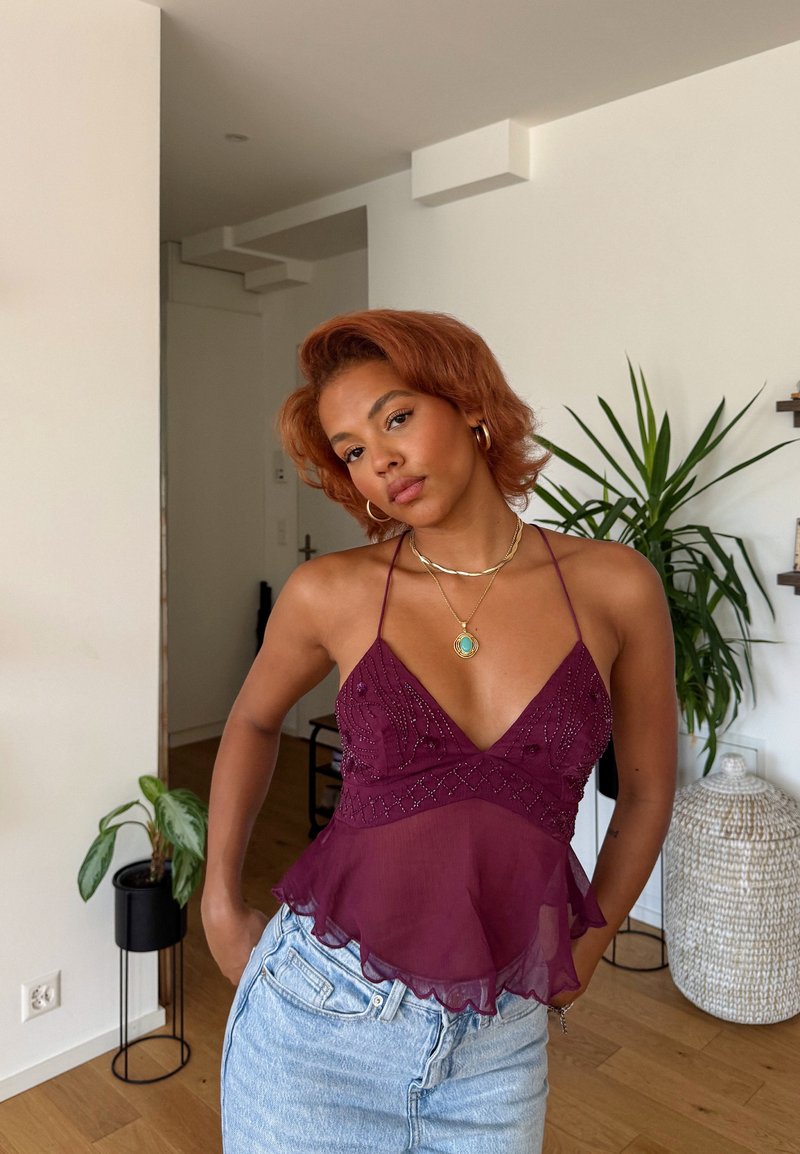 Bordeaux transparente con scollatura a V, decorata con perline e un orlo svasato; abbinata a jeans azzurri. Piante e decorazioni sullo sfondo.