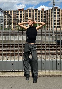 Melns piedurkņu krekls ar vertikālām līnijām, pelēkas pinstripes svārki un melni platformas apavi. Fona redzami dzelzceļa sliedes un rūpniecības ēkas.