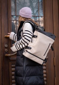 Beige batoh s rolovacím vrškem, černými akcenty, nastavitelnými popruhy a přední kapsou na zip. Nese ho osoba v černé péřové vestě a pruhovaném svetru.