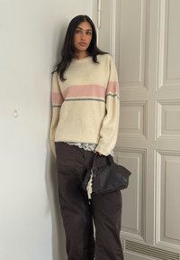 Pull crème avec des rayures roses et grises, coupe ample, associé  à un pantalon marron foncé. Petit sac à main noir avec un détail porte-clés.