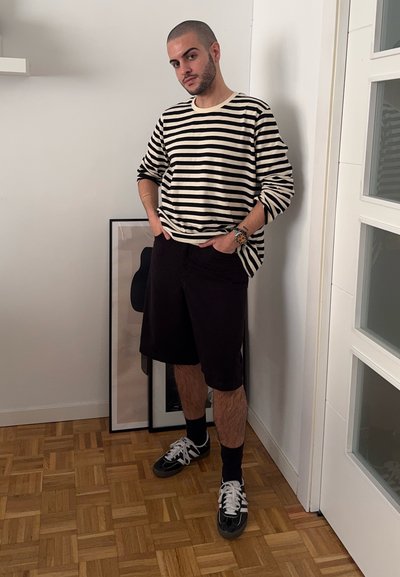 Camisa de rayas en blanco y negro de manga larga, pantalones cortos negros, calcetines tipo crew y zapatillas deportivas. El fondo incluye obras de arte enmarcadas y una puerta.