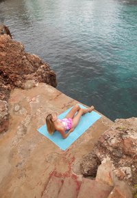 Badpak in een roze-wit bloemenpatroon, liggend op een blauwe handdoek op een rotsachtige rand bij helder water. Ruwe stenen oppervlakte en trappen zichtbaar.