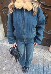 Denimist jakk kunstkarusnahaga, sobitatud sarnaste denimpükstega. Must käekott lühikeste rihmadega, terava ninaga mustad jalanõud. Tekstuurse pinnaga kõnnitee taust.