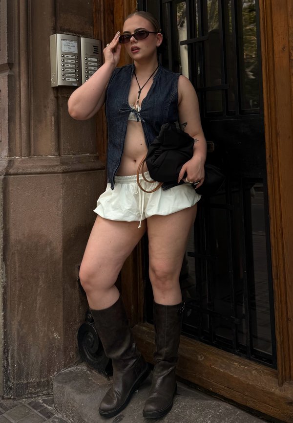 Jeansweste, weißer Minirock und knielange braune Lederstiefel. Accessoire umfasst eine schwarze Tasche. Steht neben einer Holztür mit Gegensprechanlage.