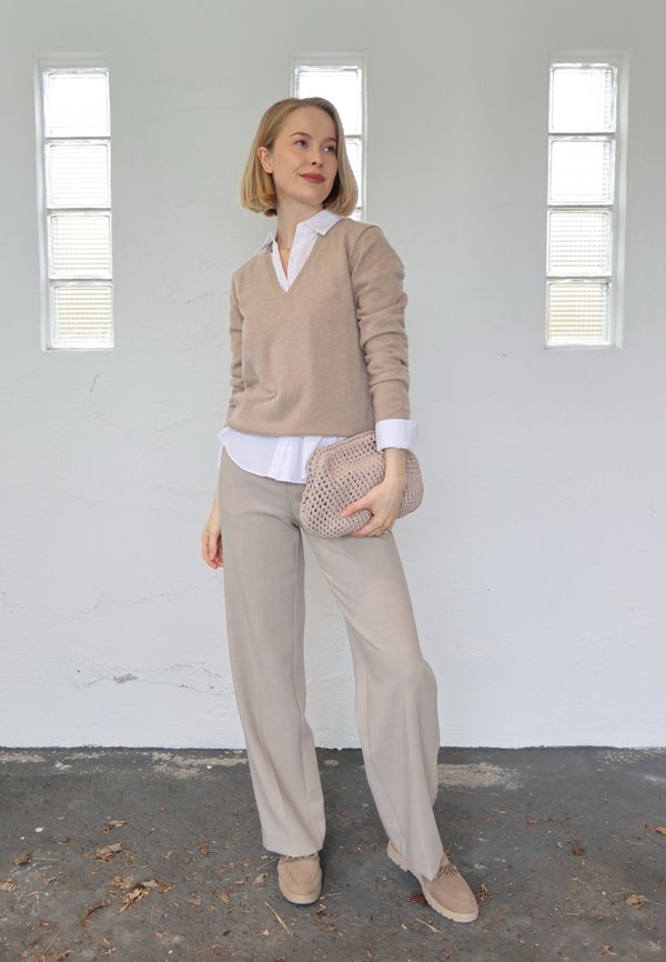 Woman standing indoors wearing beige sweater, white shirt, beige pants, holding a knitted beige clutch, with three tall windows behind her.