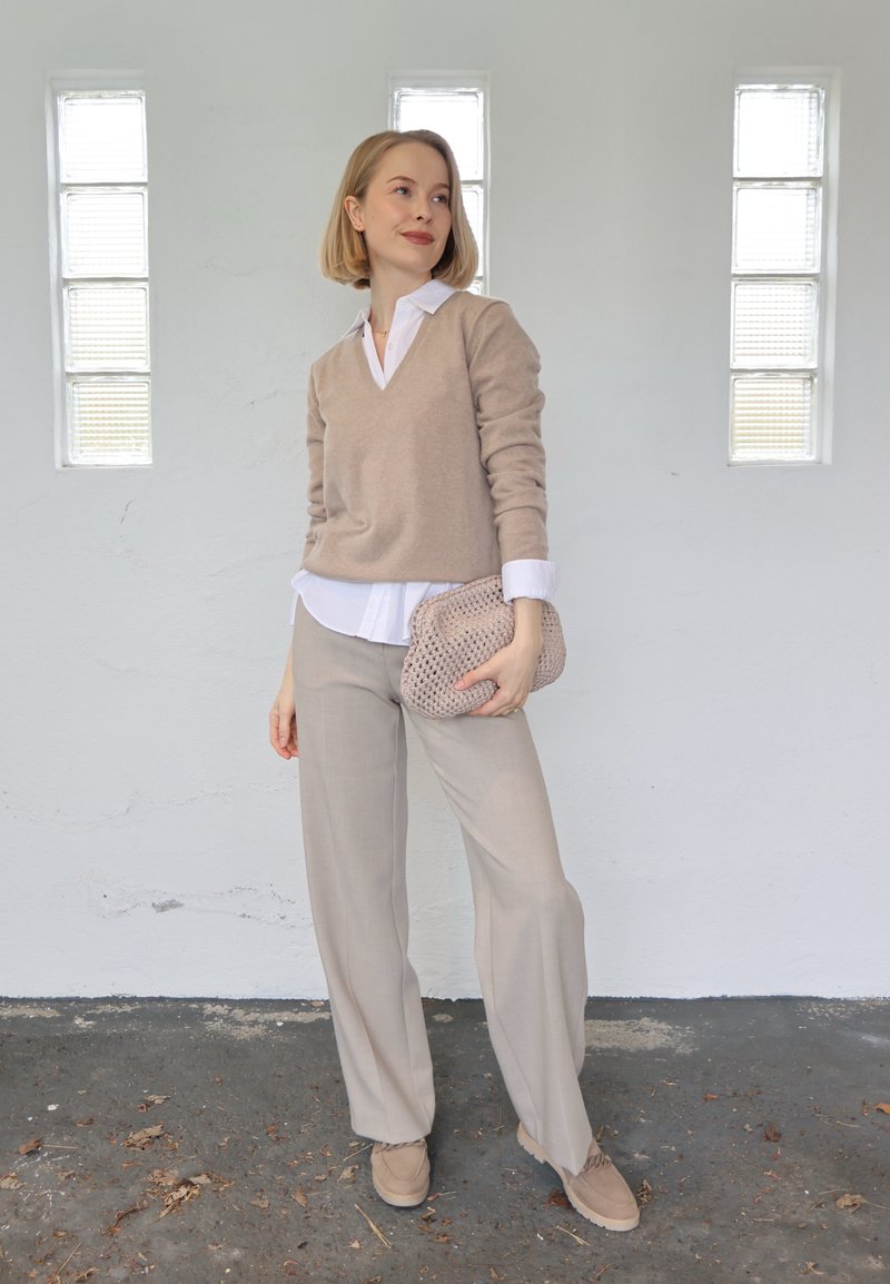 Femme debout à l'intérieur portant un pull beige, une chemise blanche, un pantalon beige, tenant une pochette beige tricotée, avec trois grandes fenêtres derrière elle.