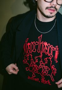 Man wearing glasses, black coat, black T-shirt with red graphic of three lions and text "Detroit, Mich." in gothic font.