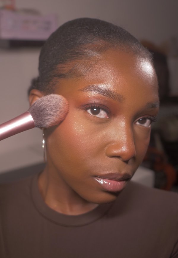 Pinceau de maquillage à manche rose appliquant du blush sur un teint foncé et lisse. La personne a les cheveux courts, porte une chemise marron et des boucles d'oreilles en créole.