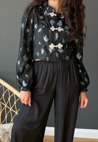 Schwarze, kurz geschnittene Bluse mit floralen Stickereien, Rüschensaum und silbernen Toggle-Verschlüssen. Kombiniert mit lockeren schwarzen Hosen. Weicher Stofftexture.