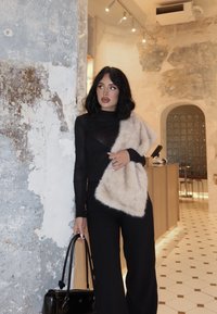 Mujer de cabello negro vestida con un conjunto negro y un estola de piel beige, sosteniendo un bolso negro, de pie contra una pared texturizada en una habitación moderna.