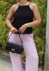 Μαύρο αμάνικο τοπ με λεπτές τιράντες, σε συνδυασμό με ανοιχτό ροζ ριγέ παντελόνια σε φαρδιά γραμμή. Ένα μαύρο τσαντάκι με χρυσές λεπτομέρειες κρατιέται στο χέρι.