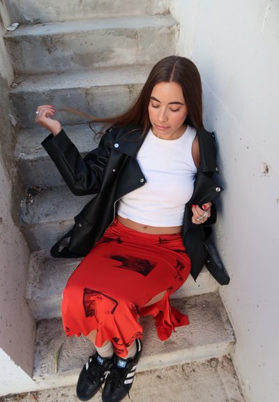 Chaqueta de biker de cuero negro sobre un top corto blanco, falda roja con patrones abstractos en negro y zapatillas negras con acentos blancos sobre escalones de concreto.