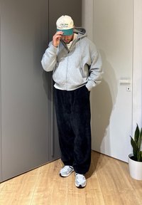 Homme en sweat à capuche gris, pantalon en velours côtelé noir, baskets blanches et casquette blanche-verte touchant la visière, debout à l'intérieur près d'une plante et d'une porte fermée.