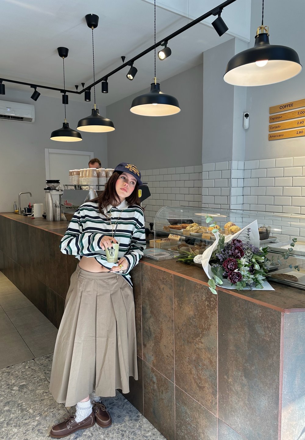 Jeune femme en chemise rayée et longue jupe s'appuyant sur le comptoir d'un café avec des pâtisseries, tenant une boisson, avec des fleurs sur le comptoir.
