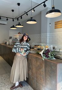 Jeune femme en chemise rayée et longue jupe s'appuyant sur le comptoir d'un café avec des pâtisseries, tenant une boisson, avec des fleurs sur le comptoir.