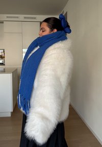 Fehér műszőrme kabát széles ujjakkal, kék sállal párosítva. A sál rojtos részletekkel és sima textúrával rendelkezik. Egyszerű fekete szoknya.