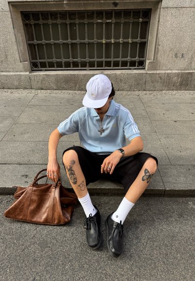 Camisa polo a rayas azul claro, pantalones cortos negros, calcetas blancas, zapatos negros, bolso de cuero marrón. Tatuajes visibles en las piernas. Gorro de béisbol blanco puesto.