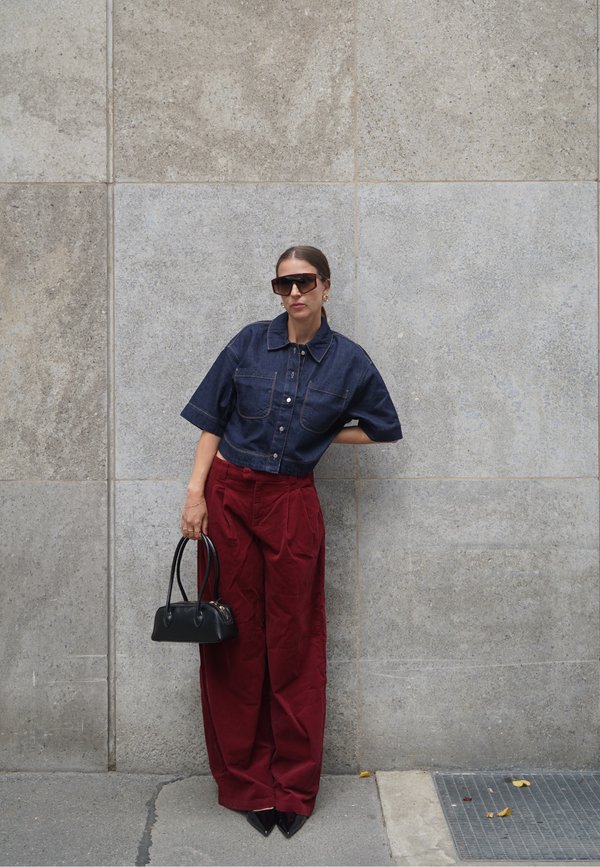 Woman wearing dark denim cropped shirt, wide red pants, black pointed shoes, oversized sunglasses, holding black handbag, leaning against stone wall.