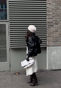 Schwarze Lederjacke, weißes Spitzenkleid, braune Stiefeletten, Pelzmütze, hält weiße Tasche mit schwarzen Punkten, steht vor einer grauen Wand.