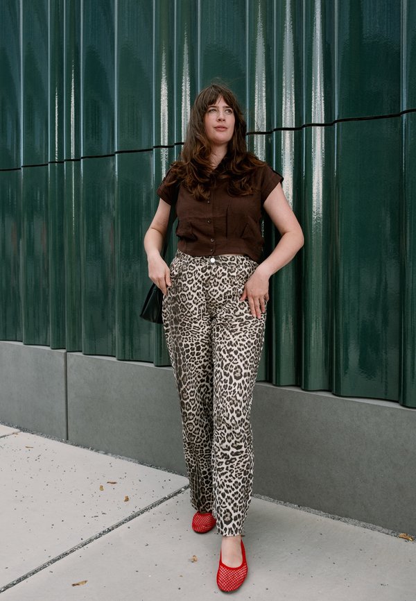 Mujer con cabello largo y castaño, vestida con una camisa marrón, pantalones con estampado de leopardo y zapatos rojos, de pie contra una pared de metal ondulado verde.