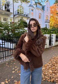 Jaqueta de camurça sintética castanha com trim fofo castanho, jeans azuis de perna larga e óculos de sol redondos. O fundo apresenta uma rua cheia de folhas e edifícios elegantes.