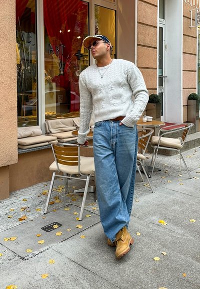 Suéter blanco de punto trenzado, jeans de corte holgado en azul claro, botas color marrón. De pie junto a una cafetería en la acera con hojas amarillas dispersas. Ventana decorativa.