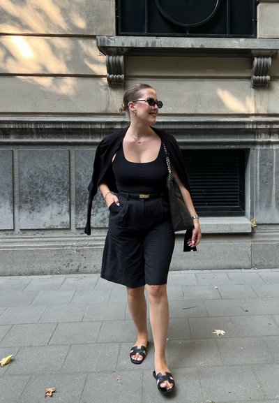 Camiseta sin mangas negra con shorts negros con cinturón y un cárdigan negro sobre los hombros. Los accesorios incluyen gafas de sol y un bolso negro.