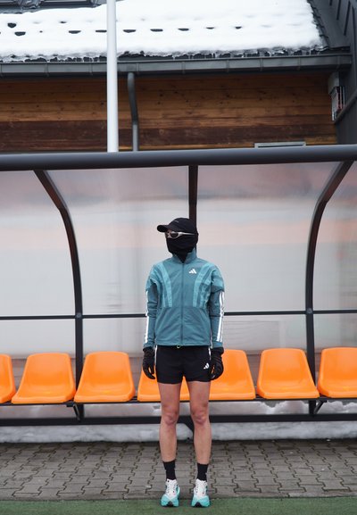 Persona con ropa deportiva, cubierta facial y gafas de sol, de pie frente a asientos naranjas bajo un refugio transparente, con nieve en el suelo y en el techo.