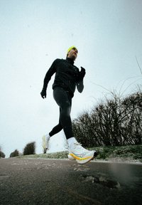 Coureur en tenue de sport noire et chapeau jaune jogging dehors par une journée enneigée près de buissons sans feuilles sur un chemin pavé.