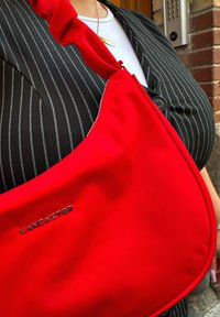 Mala de tecido vermelho com um design curvado, apresentando uma alça atada e a marca em prata "Lancaster" na frente. A textura é lisa.