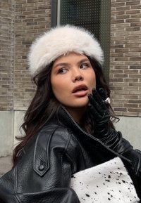 Schwarze Lederjacke, weißes Pelzstirnband und schwarze Handschuhe. Hält eine schwarz-weiß gefleckte Clutch. Neutraler Hintergrund mit Ziegelsteinen.