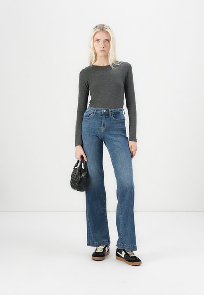 Camiseta gris de manga larga combinada con jeans acampanados azules, bolso negro y zapatillas en blanco y negro. Top de corte ajustado, denim de lavado medio, silueta contemporánea.