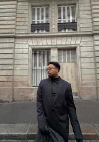 Blazer noir à la coupe moderne, porté sur un col roulé noir. Texture lisse et design ajusté. La personne tient un vêtement noir matelassé.