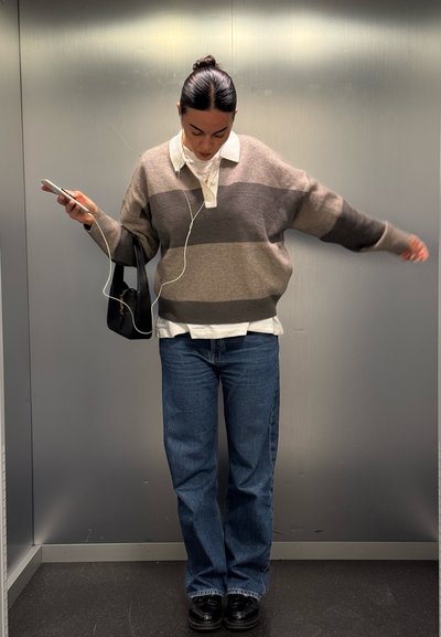 Jersey de rayas gris y marrón de talla grande sobre una camisa blanca de cuello, combinado con jeans azules y zapatos negros, sosteniendo un teléfono y una bolsa.