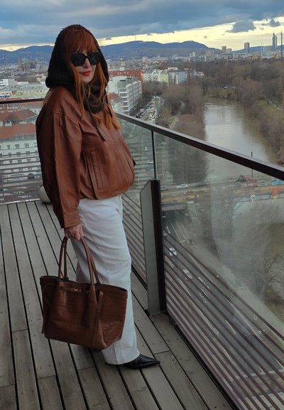 Mujer con cabello rojo, chaqueta marrón y pantalones blancos sostiene un gran bolso marrón en un balcón con vistas al río y a la ciudad bajo un cielo nublado.