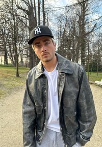 Svart läderjacka med blanka yta, stor krage och tryckknappar, bärs över en vit t-shirt; basebollkeps med logotyp.