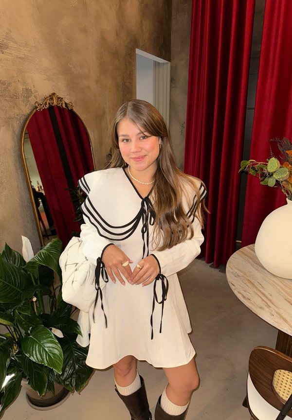 Hvit kjole med svarte detaljer og bueør, kombinert med knelengde brune støvler og en hvit veske. Bakgrunnen har røde gardiner og grøntområde.