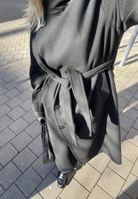 Musta, pitkä villakangastakki, jossa on vyötäröllä vyö, napillinen etupuoli ja leveät hihat. Käytetty mustien hanskojen ja kenkien kanssa, käsissä musta laukku.