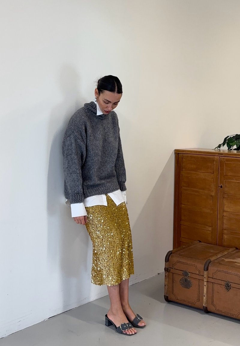 Maglione grigio a maglia, camicia bianca con bottoni sotto, gonna oro con paillettes, sandali neri a fascia. Un'unità di stoccaggio in legno sullo sfondo.