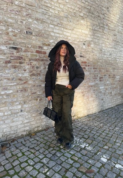 Chaqueta de plumas negra con una gran capucha, suéter crema, pantalones cargo verde olivo y un pequeño bolso negro. Fondo de adoquines texturizados.